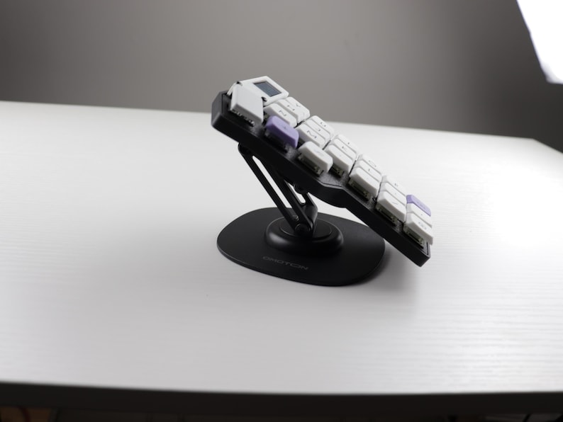 May include: A black ergonomic keyboard with white and purple keys is angled upward on a black stand. The keyboard is on a white surface, with a gray background and a bright light source.
