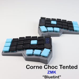 Könnte beinhalten: Zwei geteilte ergonomische Tastaturen mit schwarzen und hellblauen Tastenkappen. Die Tastaturen sind grau und haben einen kleinen Bildschirm in der Mitte. Der Text "Corne Choc Tented ZMK 'Bluetint'" befindet sich unten im Bild.