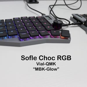 May include: A split ergonomic mechanical keyboard with black keycaps, a gray chassis, and RGB underglow lighting. The keyboard features a rotary encoder knob, a small OLED display, and is labeled Sofle Choc RGB, Vial-QMK, and MBK-Glow.
