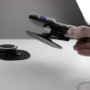 May include: Black and gray phone mount with a circular base and a pivoting arm. The mount is designed to hold a phone and is shown in use. The image also shows a separate base for the phone mount.