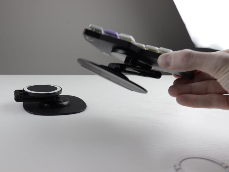 May include: Black and gray phone mount with a circular base and a pivoting arm. The mount is designed to hold a phone and is shown in use. The image also shows a separate base for the phone mount.