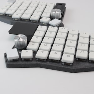 Könnte beinhalten: Eine geteilte ergonomische Tastatur mit weißen Tastenkappen und einer dunkelgrauen Basis. Die Tastatur verfügt über einen Drehgeber und eine schwarze Verbindungsbrücke. Die Tasten sind mit Buchstaben, Zahlen und Symbolen beschriftet. Die Tastatur ist auf Komfort und Effizienz ausgelegt.