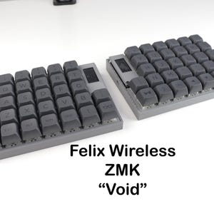 May include: A split ergonomic mechanical keyboard with dark grey keycaps and a light grey frame. The keyboard features a column-staggered layout, a small integrated display screen on each half, and the text Felix Wireless ZMK Void.
