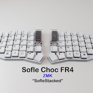 May include: A split, ergonomic keyboard with white keycaps and a white frame. The keyboard is labeled "Sofle Choc FR4" and "SofleStacked". The keys are arranged in a split layout, with a small screen on each side. The keyboard is designed for comfort and efficiency.