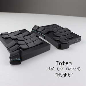 Peut inclure: Deux claviers ergonomiques noirs divisés avec des dispositions de touches incurvées. Les claviers sont étiquetés "Totem Vial-QMK (câblé) Night". Les touches sont sombres et disposées en deux parties.