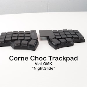 Può includere: Una tastiera Corne Choc Trackpad nera con tasti quadrati e un trackpad. La tastiera è divisa in due sezioni. Il testo "Corne Choc Trackpad", "Vial-QMK" e "NightGlide" è visualizzato sotto.