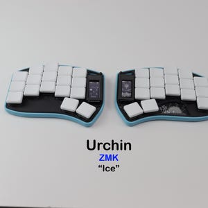 Pode incluir: Um teclado ergonómico dividido com uma moldura azul-clara e teclas brancas. O dispositivo possui dois pequenos ecrãs retangulares e uma placa de base preta com um gráfico de ouriço. O texto abaixo diz Urchin ZMK Ice.