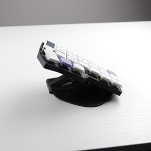 May include: A black and white ergonomic keyboard with white and purple keys is angled upwards on a black stand. The keyboard is designed for comfort and efficiency, ideal for office or home use.