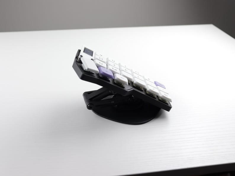 May include: A black and white ergonomic keyboard with white and purple keys is angled upwards on a black stand. The keyboard is designed for comfort and efficiency, ideal for office or home use.
