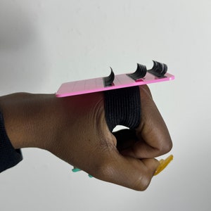 Könnte beinhalten: Rosa Wimpernverlängerungs-Tablett mit einem schwarzen Gummiband. Das Tablett hat mehrere Reihen zum Halten einzelner Wimpern.