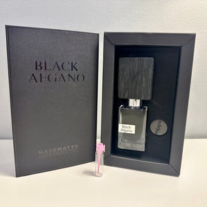 Op de afbeelding: Een zwarte doos met de tekst "Black Afgano" en het logo "Nasomatto" op de voorkant. De doos is open en een klein flesje parfum is zichtbaar in de doos. Het flesje is van helder glas met een zwarte dop en de tekst "Black Afgano" op het etiket. Een klein proefbuisje parfum is ook zichtbaar.