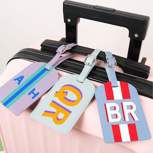 Könnte beinhalten: Drei personalisierte Gepäckanhänger in Lila-, Blau- und Hellblautönen mit weißen Nähten und silbernen Schnallen. Jeder Anhänger zeigt unterschiedliche Initialen in kräftigen, kontrastierenden Farben. Die Anhänger liegen auf einem rosa Koffer.