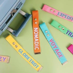 Könnte beinhalten: Eine Auswahl an farbenfrohen Gepäckanhängern mit Wörtern wie "Expedition" und "Reise". Die Anhänger sind in verschiedenen Farben wie Orange, Pink und Blau gehalten und liegen in der Nähe eines hellblauen Koffers auf grünem Untergrund.