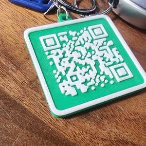 Puede incluir: Un llavero con código QR verde y blanco. El llavero cuadrado tiene un fondo verde con un diseño de código QR blanco. También se ven un llavero azul, llaveros plateados y una llave de coche.