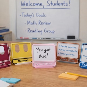 May include: A selection of colourful desk organisers with small whiteboards, each with a different message. The organisers are in shades of red, yellow, pink, and blue. A whiteboard in the background displays "Welcome, Students!" and daily goals.