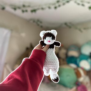 May include: A handmade crochet ferret toy. The ferret has a white body, brown head and feet, and a pink mouth. The toy is held up in front of a blurred background. The ferret is approximately 6 inches long.