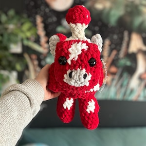 May include: A red and white crocheted plush toy with a mushroom on its head. The toy has a white star pattern and black eyes.