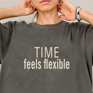 May include: A charcoal gray short-sleeved t-shirt featuring the text "TIME feels flexible" printed in a light beige, sans-serif font across the chest. The garment has a crew neckline and a relaxed fit with a slightly faded, washed-out appearance.