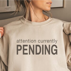 May include: A beige crewneck sweatshirt featuring the dark brown text "attention currently PENDING" printed across the chest. The garment has a ribbed collar and long sleeves, providing a neutral-toned look with clear, bold lettering.