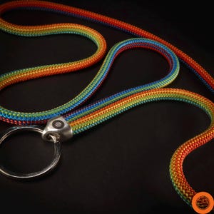Könnte beinhalten: Ein mehrfarbiges Schlüsselband mit einem silberfarbenen Schlüsselring. Das Schlüsselband besteht aus einem dicht gewebten, strapazierfähigen Material in einem Farbverlauf aus Rot, Orange, Gelb, Grün und Blau. Der Schlüsselring ist an einem silberfarbenen Metallverschluss befestigt.