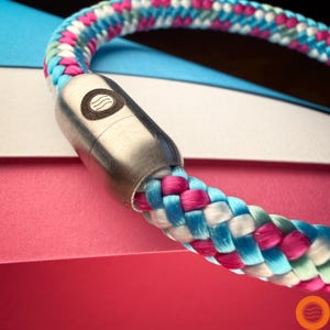 Op de afbeelding: Close-up van een kleurrijke hondenriem. De riem is gemaakt van geweven touw in roze, blauwe, witte en groene tinten. Het heeft een zilverkleurige metalen sluiting met een cirkelvormig ontwerp.