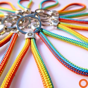 Könnte beinhalten: Eine Sammlung von Regenbogen-Schlüsselanhängern mit silbernen Metallringen. Jeder Schlüsselanhänger hat ein geflochtenes Band in leuchtenden Farben von Rot, Orange, Gelb, Grün und Blau. Die Schlüsselanhänger sind kreisförmig angeordnet und zeigen ihr farbenfrohes Design.