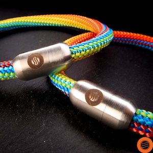 Pode incluir: Um cordão de corda trançada com um padrão de cores arco-íris vibrantes em vermelho, laranja, amarelo, verde e azul. O cordão é preso por dois fechos cilíndricos de metal prateado escovado, cada um exibindo um logotipo circular com três linhas onduladas.