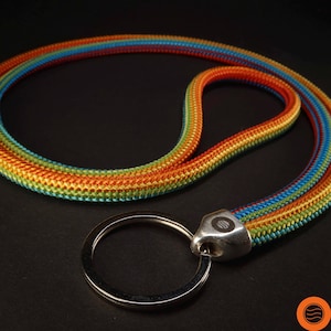 Puede incluir: Un cordón de arcoíris con un llavero plateado. El cordón está hecho de cuerda trenzada y tiene un patrón colorido de rojo, naranja, amarillo, verde, azul y morado.