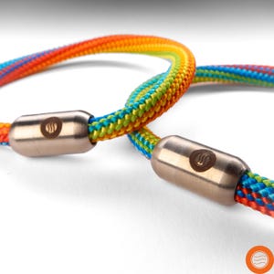 Könnte beinhalten: Zwei bunte Armbänder aus Seil mit Metallverschlüssen. Die Armbänder bestehen aus einem regenbogenfarbenen Seil mit einem Muster aus Rot, Orange, Gelb, Grün, Blau und Lila. Die Metallverschlüsse sind silberfarben und haben ein kleines Logo.
