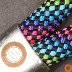 Könnte beinhalten: Eine geflochtene Kordel mit einem mehrfarbigen Muster aus schwarzen, blauen, violetten, pinken und lindgrünen Kunstfasern. Eine silberne Metallendkappe ist an der Kordel befestigt und zeigt eine kreisförmige beigefarbene Einlage mit einem weißen Drei-Wellen-Symbol.
