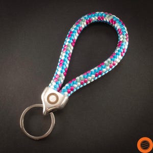 Könnte beinhalten: Ein farbenfrohes Schlüsselband mit geflochtenem Seildesign. Das Band weist eine Mischung aus blauen, rosa und silbernen Fäden auf, die an einem silberfarbenen Metallverschluss und Schlüsselring befestigt sind. Der Hintergrund ist einfarbig schwarz.
