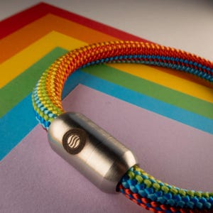 Könnte beinhalten: Eine bunte Seilkette mit einem Edelstahlverschluss. Das Seil ist eine lebendige Mischung aus Regenbogenfarben, darunter Rot, Orange, Gelb, Grün, Blau und Lila. Der Verschluss hat ein kreisförmiges Design mit einem Wellenmuster.