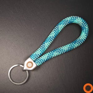 Könnte beinhalten: Ein Schlüsselanhänger aus blauem und türkisfarbenem Seil mit einem silbernen Schlüsselring. Das Seil ist geschlungen und an einem Metallstück mit einem Logo befestigt. Der Hintergrund ist dunkelgrau.