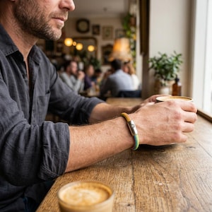 Peut inclure: Bracelet multicolore avec un motif de corde jaune, vert et bleu et un fermoir argenté. La personne porte le bracelet et tient une tasse de café. L'arrière-plan est un café avec une table en bois.