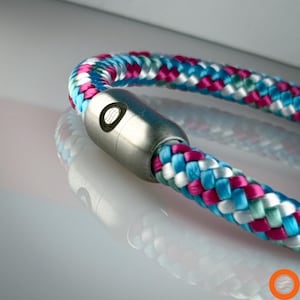 Könnte beinhalten: Nahaufnahme eines Armbands mit einem geflochtenen Seildesign in Blau-, Rosa- und Weißtönen. Das Armband hat einen silberfarbenen zylindrischen Verschluss mit einem kreisförmigen Design. Das Seil ist eng geflochten und erzeugt ein strukturiertes Aussehen.