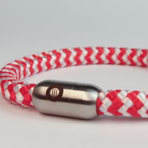 Pode incluir: Um close-up de uma pulseira de corda trançada vermelha e branca com um fecho prateado. O fecho tem um pequeno desenho circular gravado. A pulseira é feita de um material de corda espessa e durável.