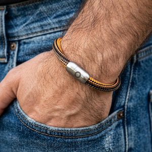 Op de afbeelding: Een gevlochten armband met een zwart, oranje en bruin patroon. De armband heeft een zilverkleurige magnetische sluiting. De armband wordt om een pols gedragen, met een blauwe jeanszak op de achtergrond.