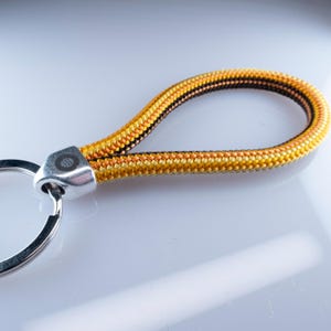 May include: A close-up of a keychain with a loop made of yellow, orange, and black braided rope. The rope is attached to a silver-colored metal piece with a small circular design. A silver key ring is attached to the metal piece.