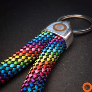 Op de afbeelding: Close-up van twee regenboogkleurige sleutelhangers met een geweven patroon. De sleutelhangers hebben een levendige reeks kleuren, waaronder rood, oranje, geel, groen, blauw en paars, tegen een zwarte achtergrond. Ze zijn bevestigd aan een zilveren metalen ring.
