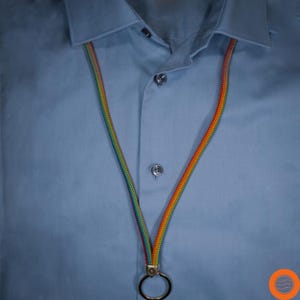 Könnte beinhalten: Ein mehrfarbiges Schlüsselband mit einem Metallring hängt vor einem hellblauen Button-Down-Hemd. Das Schlüsselband besteht aus geflochtenem Kordel in einem Farbspektrum, einschließlich Rot, Orange, Gelb, Grün und Blau. Das Hemd hat einen klassischen Kragen und Knopfverschluss.