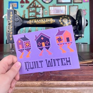 May include: A purple sticker featuring three whimsical house designs with orange and purple accents, each with legs. The text "QUILT WITCH" is printed below the houses. A vintage black sewing machine is in the background.