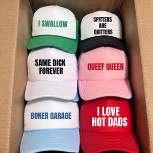 May include: Six trucker hats arranged in a cardboard box. The hats feature white front panels with various text: I SWALLOW in green, SPITTERS ARE QUITTERS in black, SAME DICK FOREVER in black, QUEEF QUEEN in red, BONER GARAGE in blue, and I LOVE HOT DADS in white.