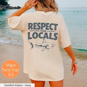 May include: A white t-shirt with a graphic of a shark and the text "Respect the Locals" printed on the back. The shirt is being worn by a person standing on a beach.