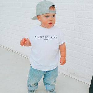 May include: A white t-shirt with the text "RING SECURITY Noah" printed on the front. A young child is wearing the shirt with a gray baseball cap.