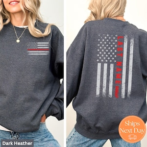 May include: Dark heather grey sweatshirt with a distressed American flag design on the back. The flag has a red stripe down the center with the text "LAST NAME" printed vertically on the stripe. The front of the sweatshirt has a smaller version of the flag design.
