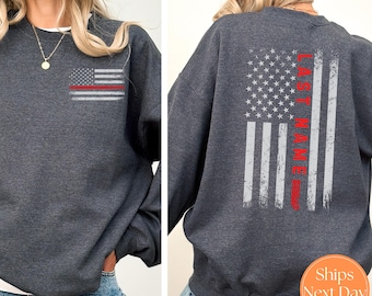 May include: Dark heather grey sweatshirt with a distressed American flag design on the back. The flag has a red stripe down the center with the text "LAST NAME" printed vertically on the stripe. The front of the sweatshirt has a smaller version of the flag design.