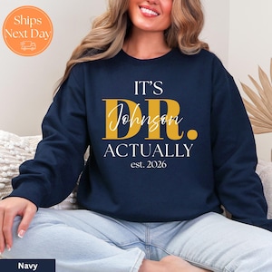 May include: Navy blue sweatshirt with the text "IT'S DR. Johnson ACTUALLY est. 2026" in gold and white lettering. The sweatshirt is a crew neck style.