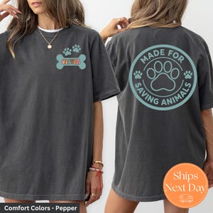 May include: Dark grey t-shirt with a front graphic of paw prints and the name "Olivia" on a bone. The back of the shirt features a circular design with paw prints and the text "Made for Saving Animals".