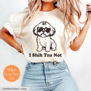 May include: Ivory-colored t-shirt featuring a black graphic of a Shih Tzu dog wearing sunglasses with the text "I Shih Tzu Not". The shirt is made of a soft, comfortable material.