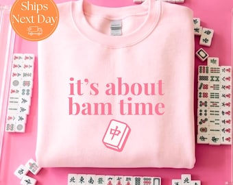 May include: A light pink crewneck sweatshirt featuring the dark pink text it's about bam time and a graphic of a mahjong tile with a red character. The garment is surrounded by various white mahjong tiles on a bright pink background.